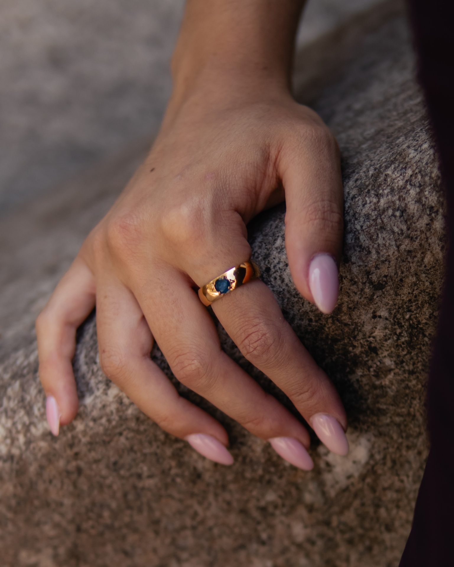 Anel dourado com pedra azul em mão com unhas rosa claro sobre pedra