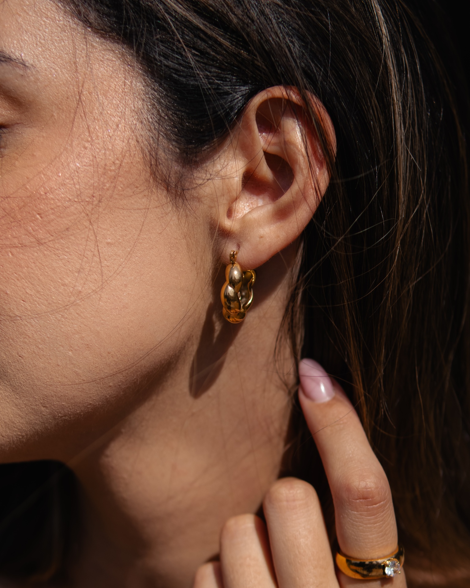 Brincos de argola dourados com design de bolhas numa orelha de mulher de cabelo castanho escuro e mão com anel dourado