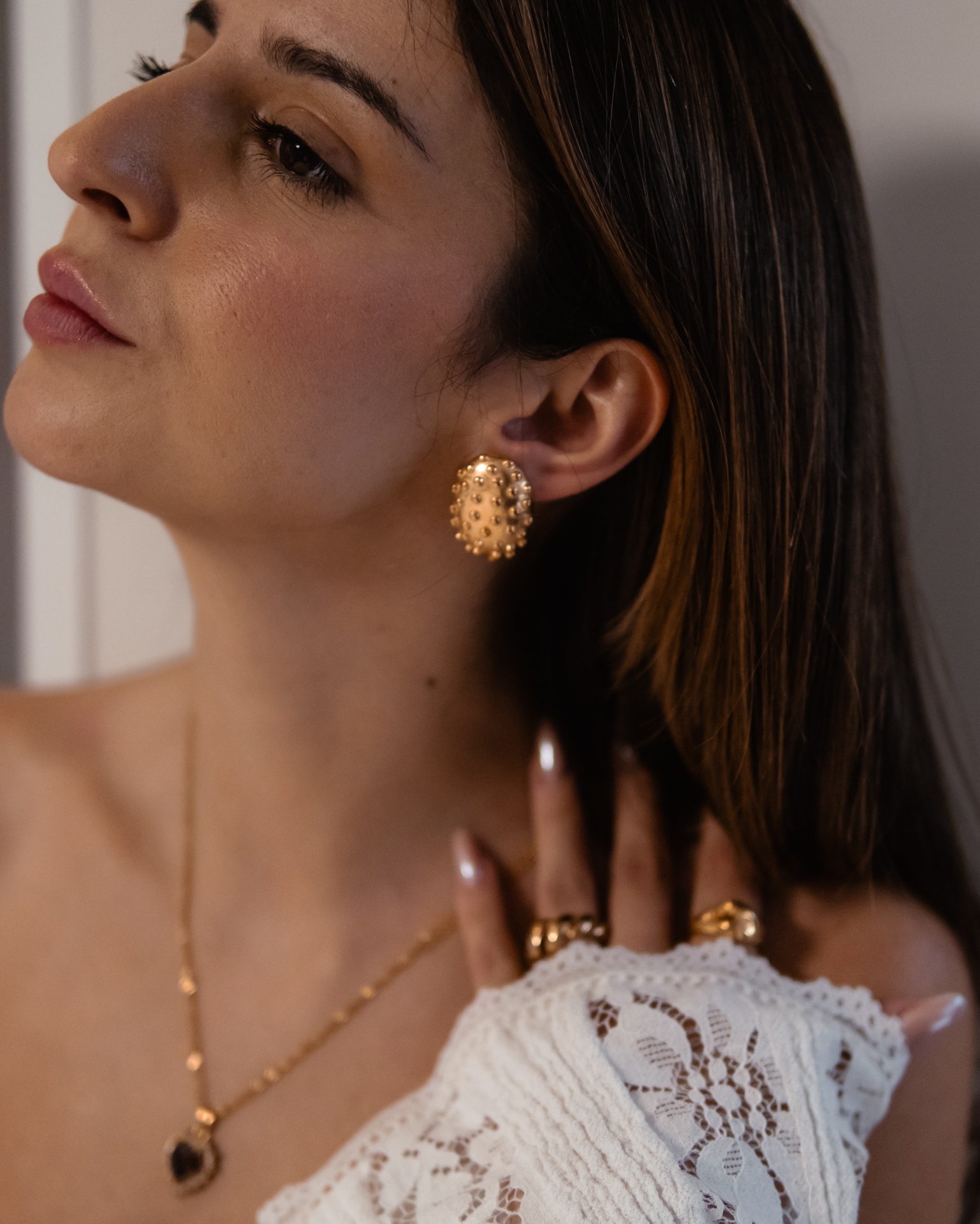 Brincos dourados ovais com pedras, colar e anéis dourados em mulher com blusa branca de renda