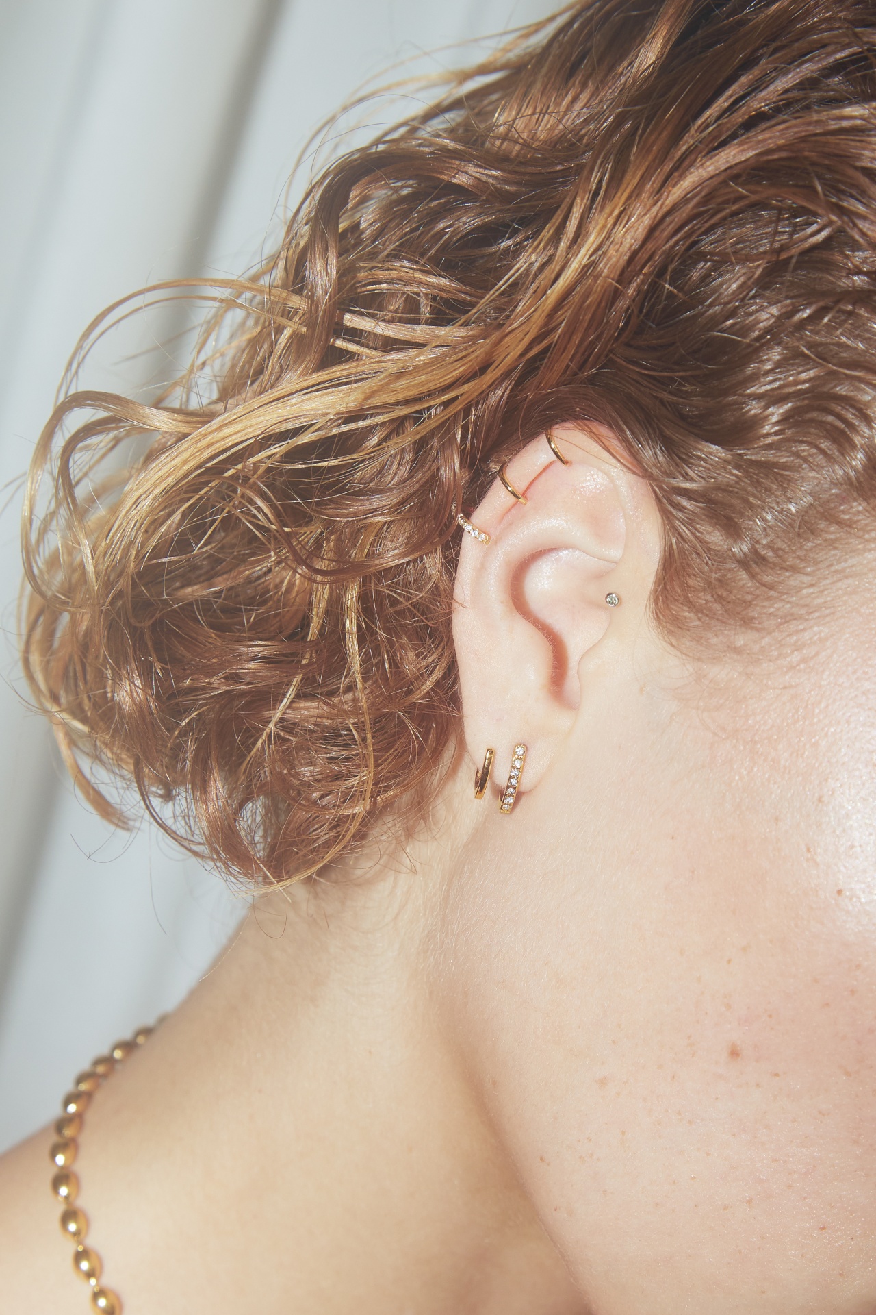 Orelha com vários brincos dourados e cabelo encaracolado preso para trás.