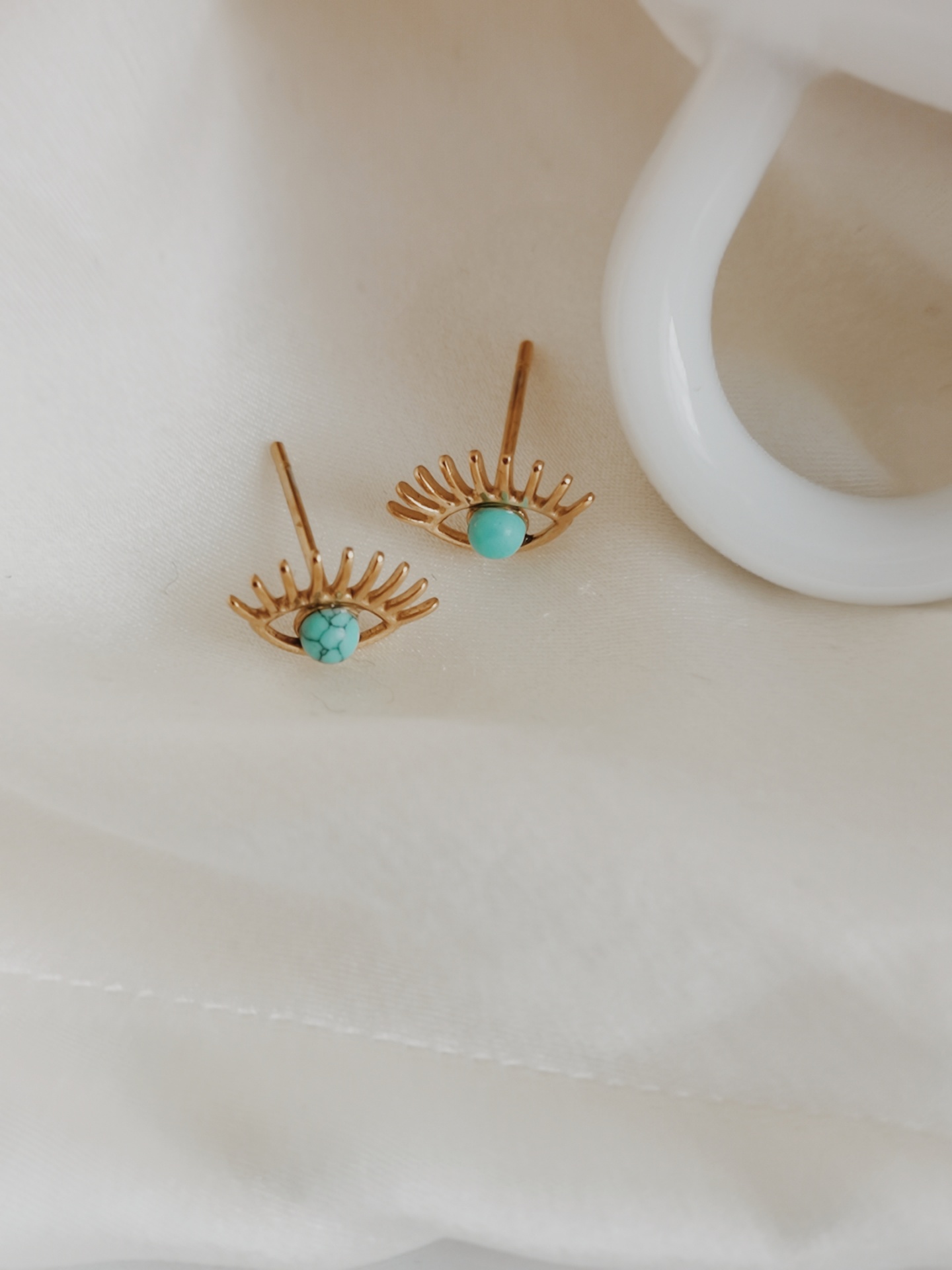 Brincos dourados em forma de olhos com pedra turquesa sobre fundo branco