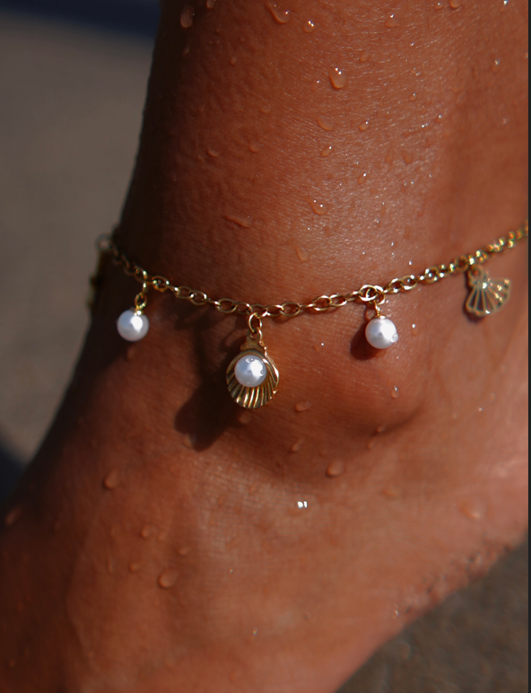 Tornozeleira dourada com pingentes de concha e pérolas num tornozelo molhado