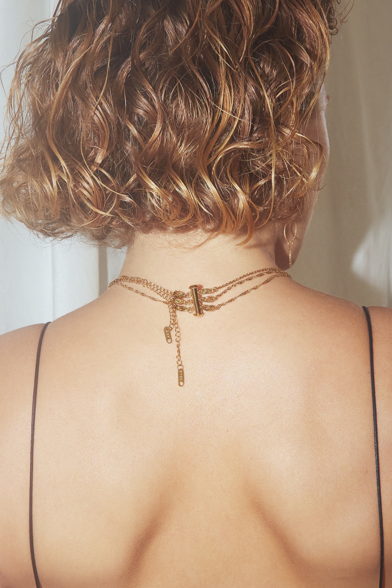 Colar dourado de três correntes finas com fecho no pescoço de pessoa de cabelo castanho encaracolado