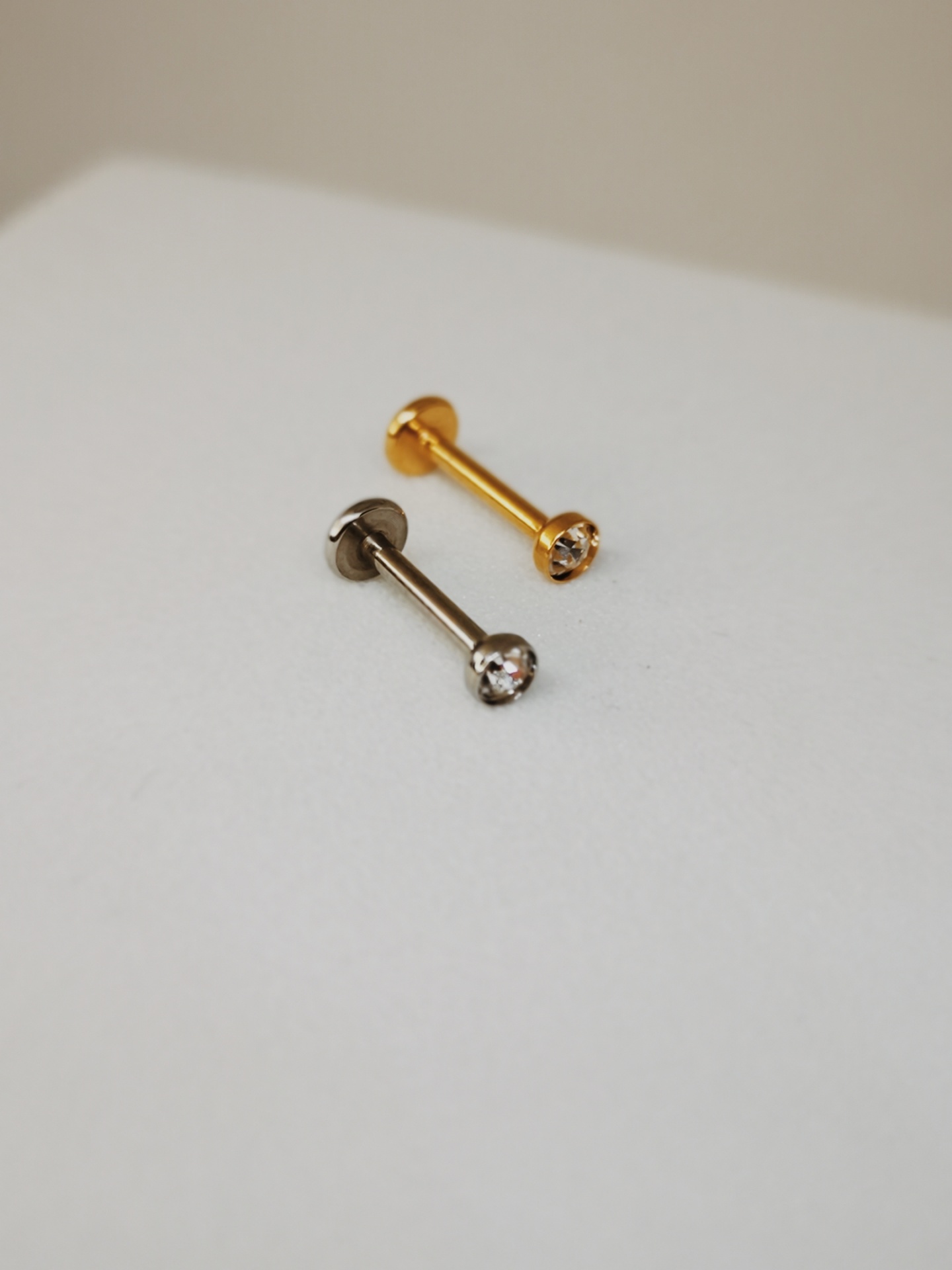 Dois brincos barra, um dourado e outro prateado, com pedras claras sobre superfície branca