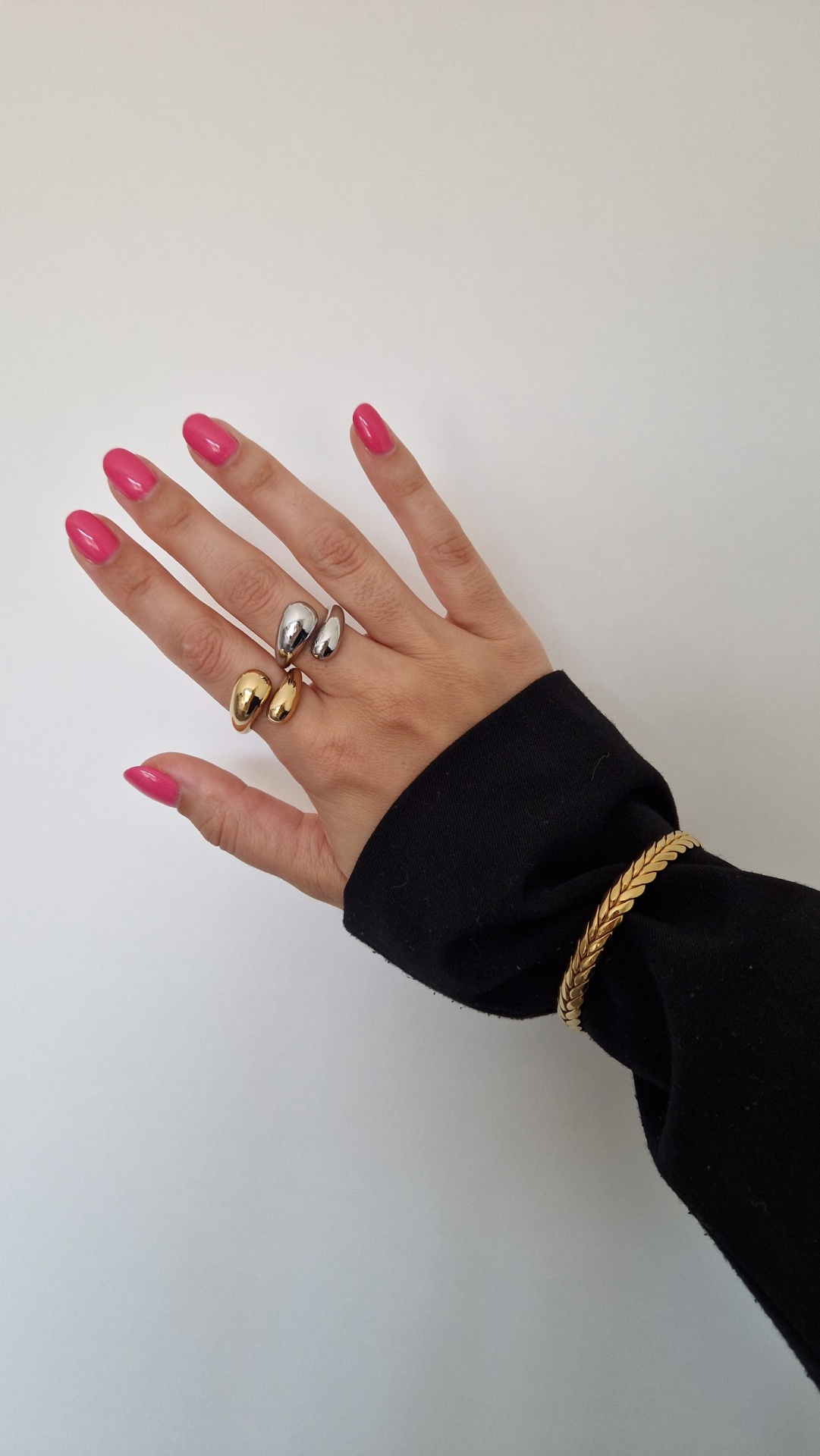 Mão feminina com unhas rosa, anéis dourado e prateado, pulseira dourada, fundo branco