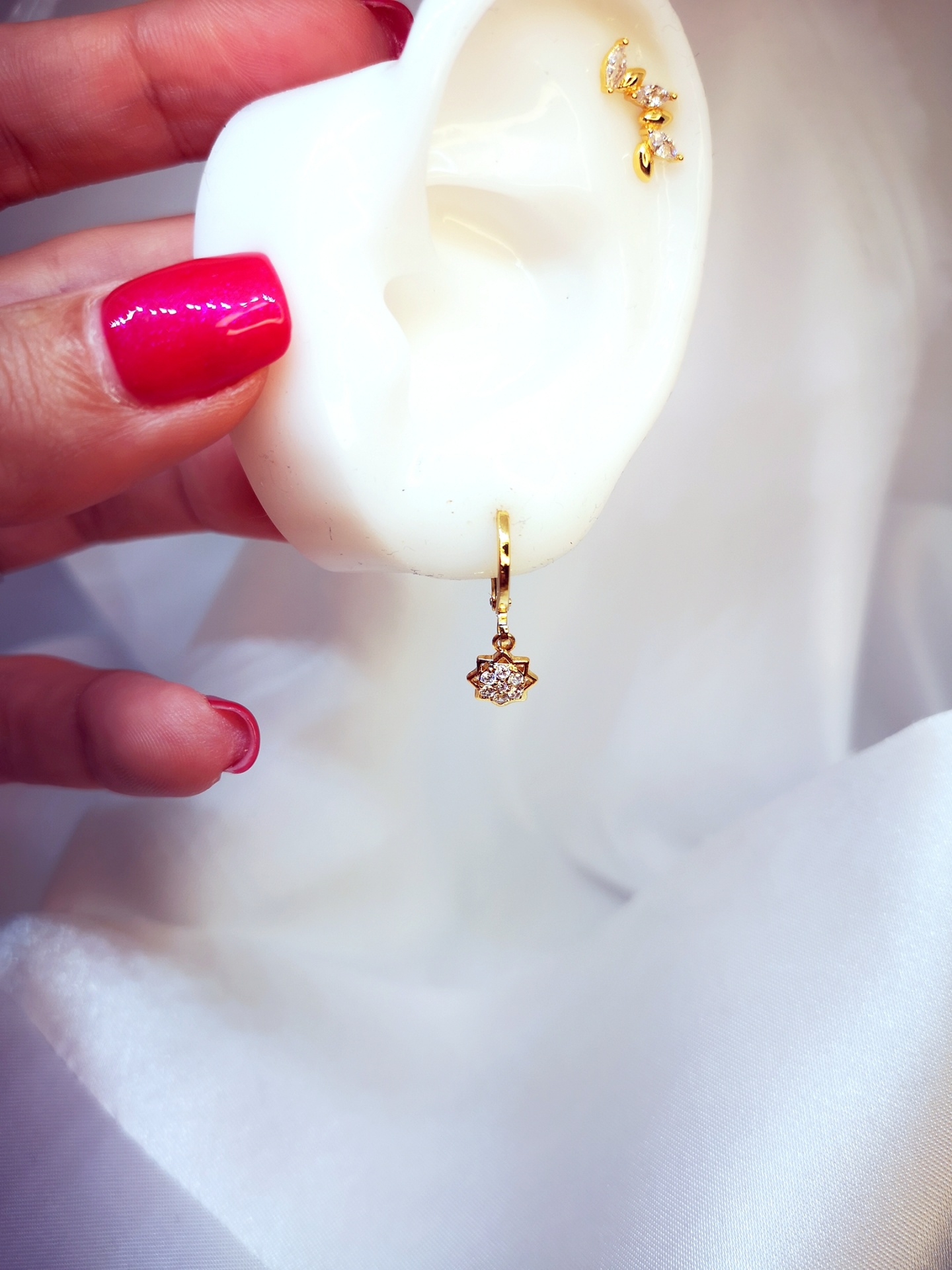 Brincos dourados com cristais em modelo de orelha branca a ser segurada com mão de unhas vermelhas.