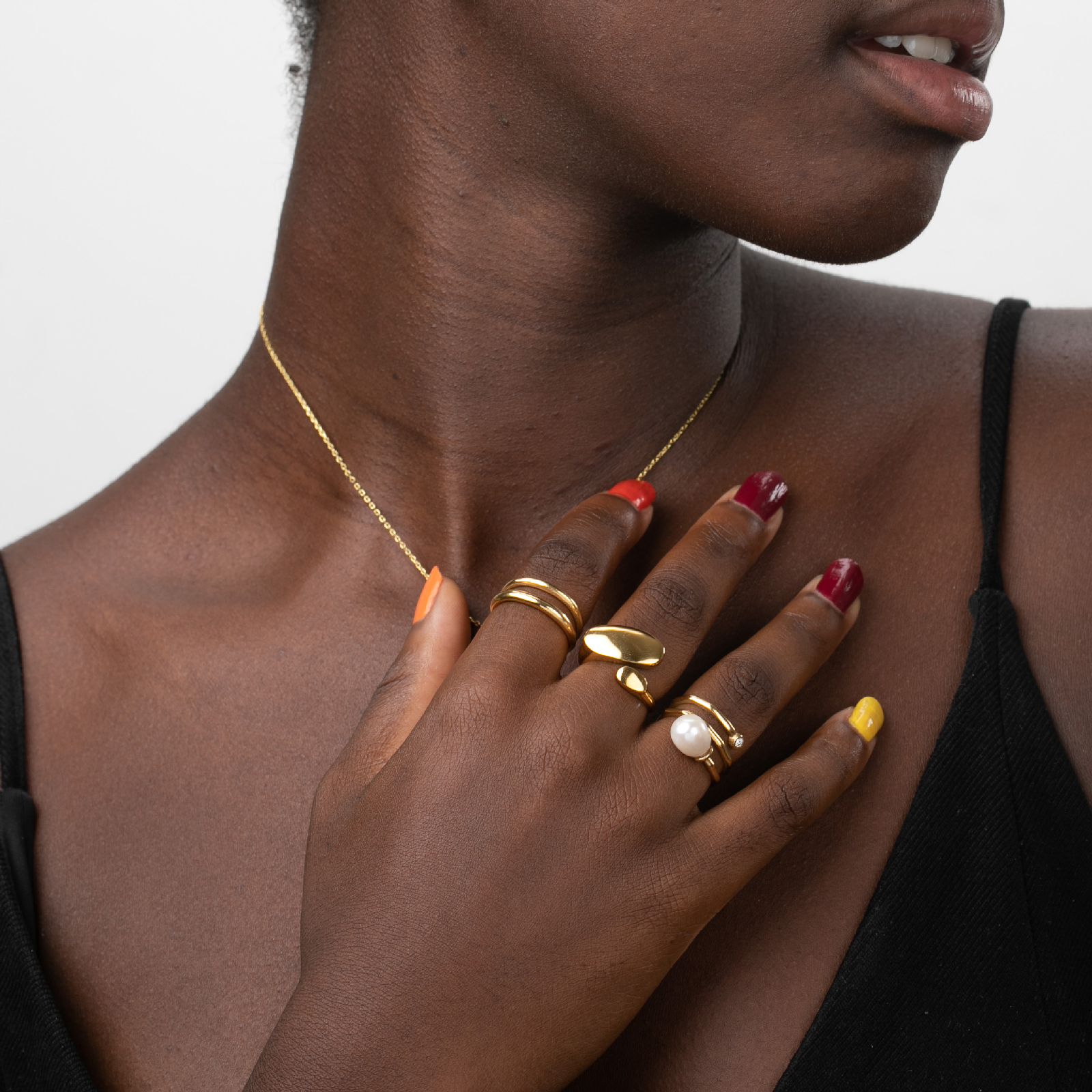 Colar dourado fino e três anéis dourados com pérola em mão com unhas coloridas e pele escura