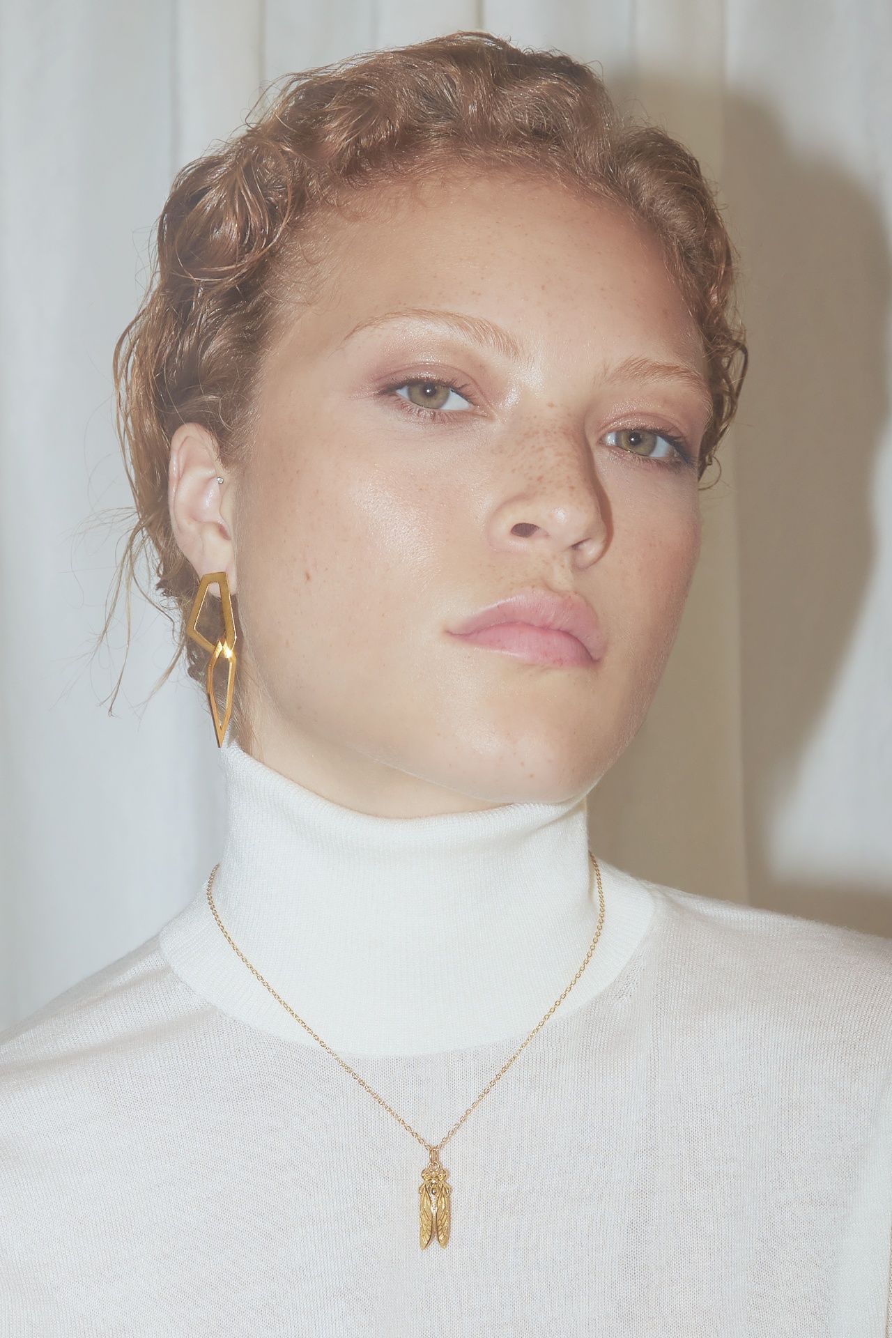 Mulher com brincos dourados e colar dourado com pendentes usando blusa branca de gola alta