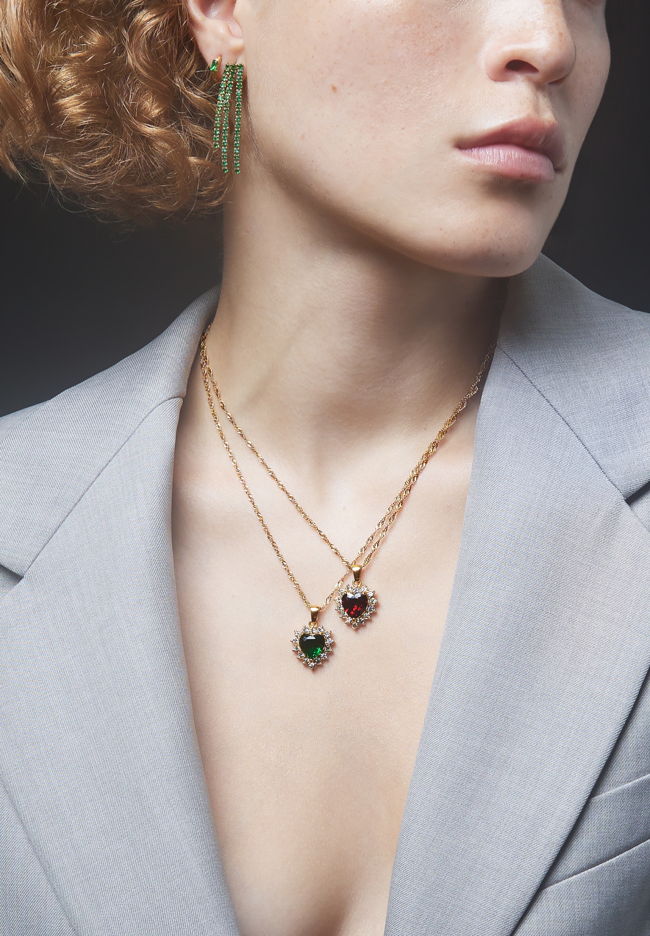 Colares dourados com pendentes em coração com pedras verde e vermelha e brincos verdes em pessoa com blazer cinzento