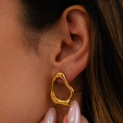 Brinco de ouro irregular usado na orelha de uma mulher com unhas peroladas.