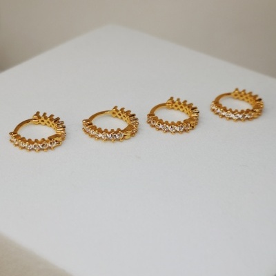 Quatro argolas douradas com pedras claras e detalhes em forma de picos sobre superfície branca