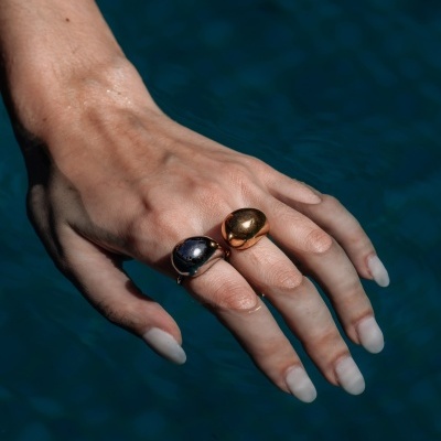 Mão feminina com dois anéis largos dourado e prateado sobre fundo azul de água.