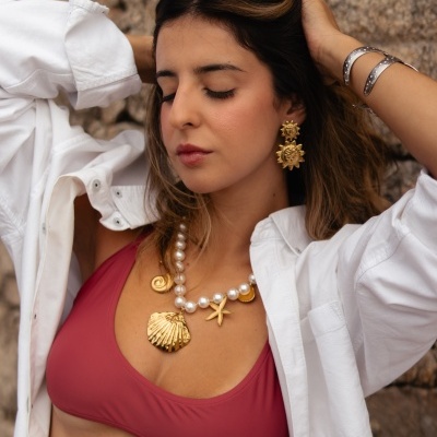 Mulher com colar e brincos dourados, pulseiras, top vermelho e camisa branca em fundo de pedra