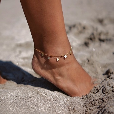 Tornozeleira dourada com pendentes de flores numa perna na areia da praia