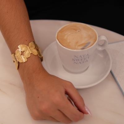Pulseira dourada de folhas em metal e chávena branca de café com leite numa mesa de mármore