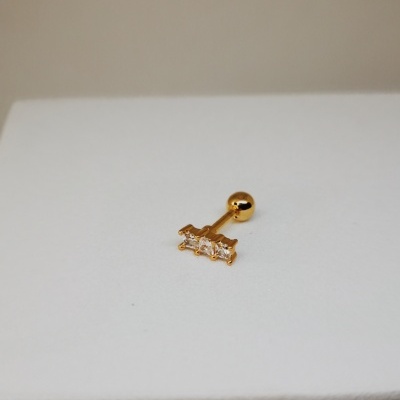 Piercing dourado com três pedras retangulares sobre superfície branca