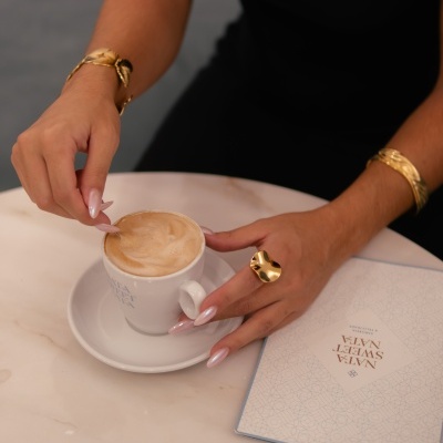 Pessoa com acessórios dourados segurando chávena branca de café com espuma sobre mesa de mármore junto a caderno com capa escrita NATA SWEET NAPA