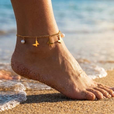 Pulseira dourada com pendentes em forma de borboleta e pérolas no tornozelo de um pé na praia