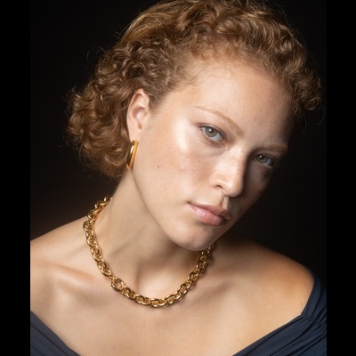 Mulher com colar e brincos dourados, cabelo curto encaracolado, fundo preto
