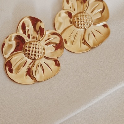 Brincos dourados em forma de flores com textura no centro, sobre tecido branco.