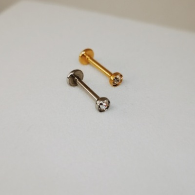 Dois brincos barra, um dourado e outro prateado, com pedras claras sobre superfície branca