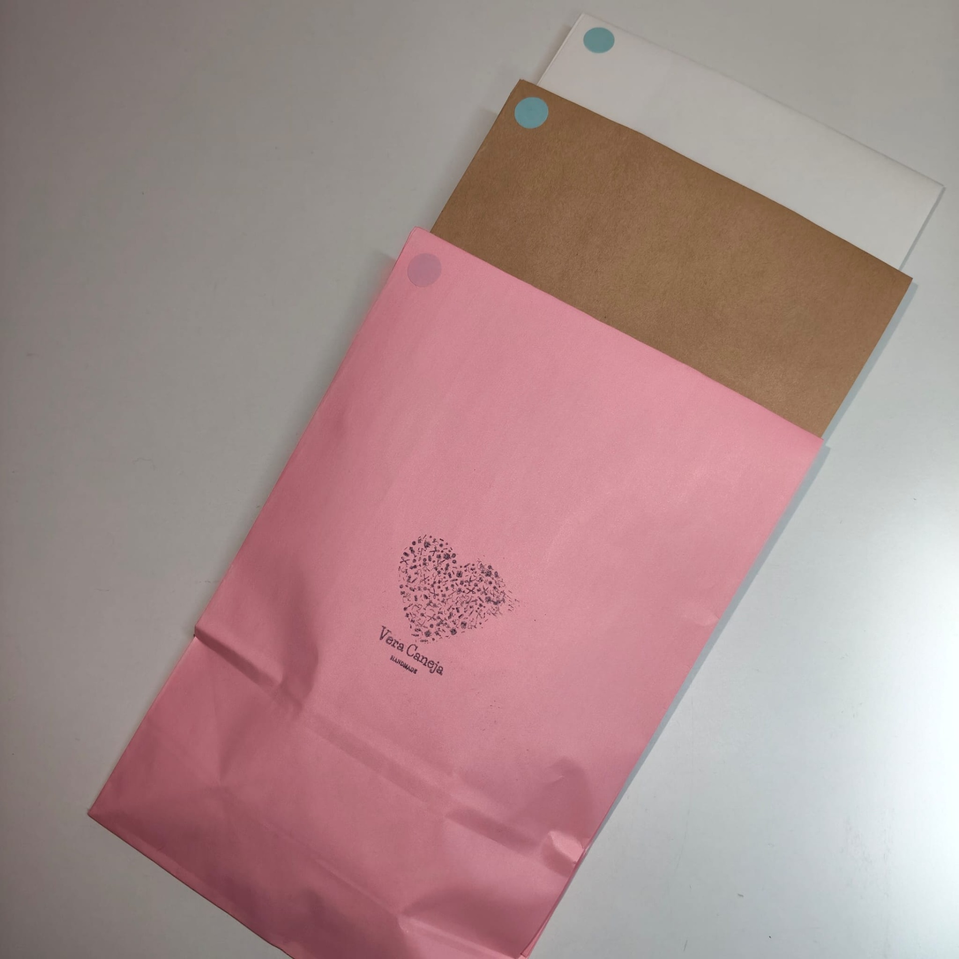 Três envelopes de papel branco, castanho e rosa com autocolantes coloridos e texto Vera Caneja HANDMADE no envelope rosa.