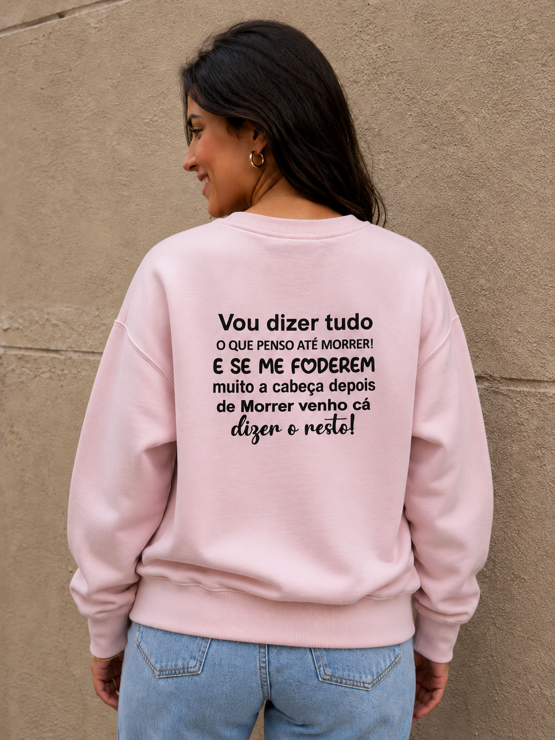 Pessoa com sweatshirt rosa claro com texto em preto nas costas, jeans azuis claros e parede bege de fundo.