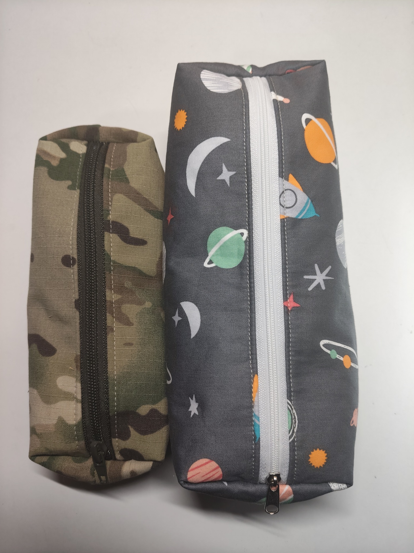 Duas bolsas retangulares, uma camuflada e outra com padrão espacial, fechadas com zíper