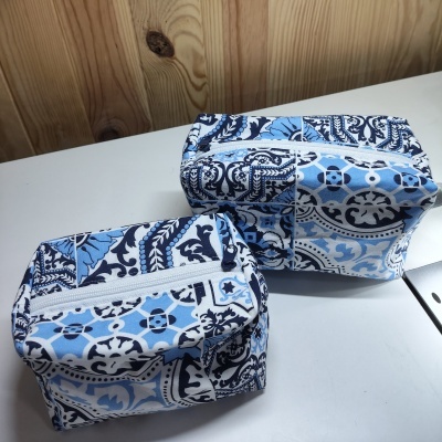 Dois necessaires com padrão azul, branco e preto com fecho branco, sobre superfície branca e fundo de madeira clara.