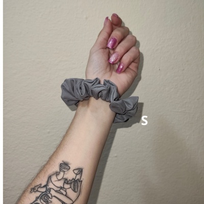 Mão com unhas glitter rosa, scrunchie cinzento e tatuagem no pulso