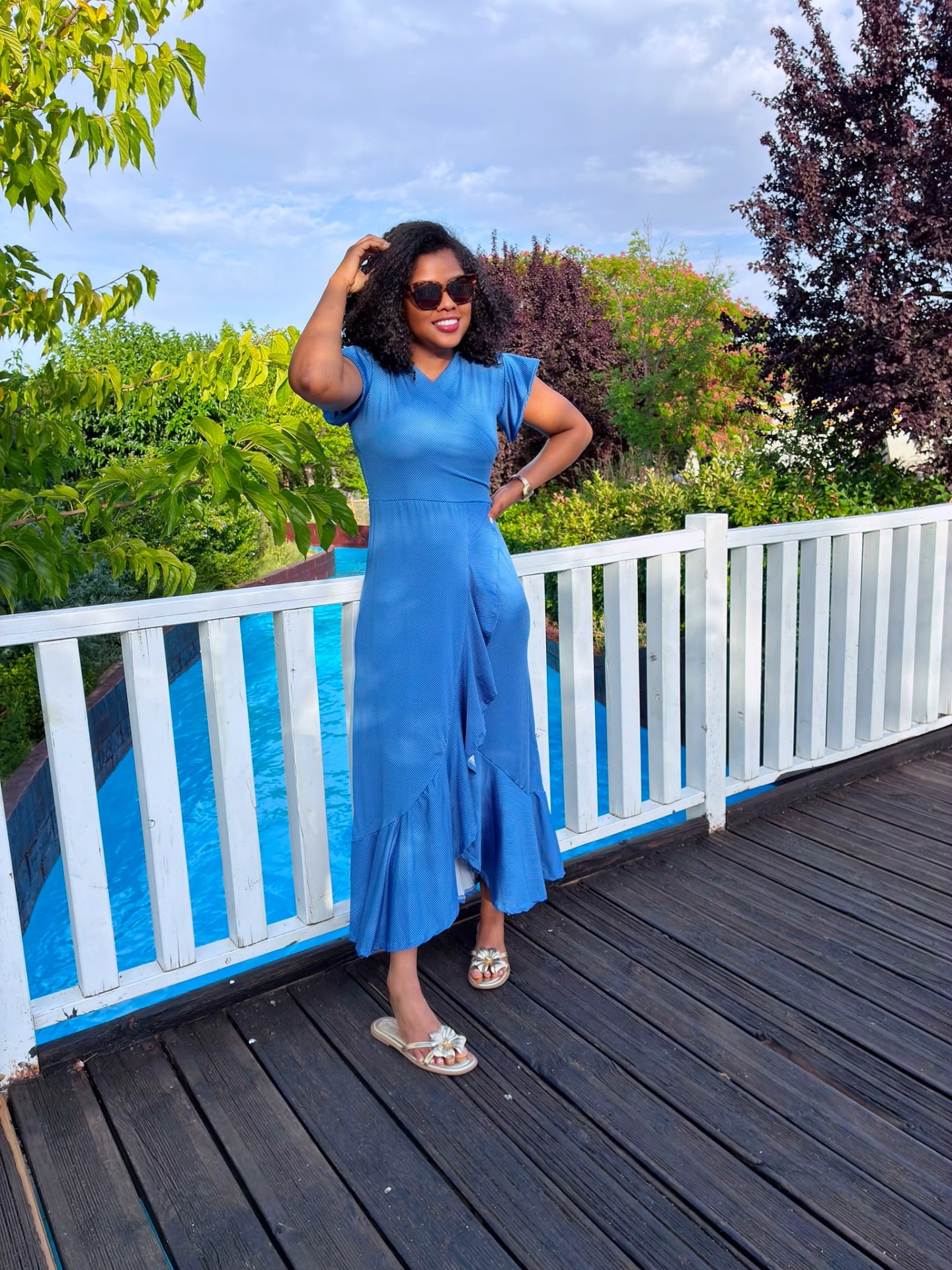 Mulher com vestido azul comprido com folho, óculos de sol e sandálias douradas de pé em passadiço de madeira junto a piscina e vegetação