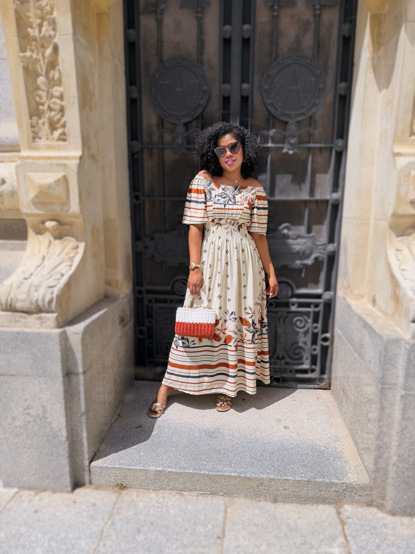 Mulher com vestido longo floral e riscas, mala trançada e sandálias douradas