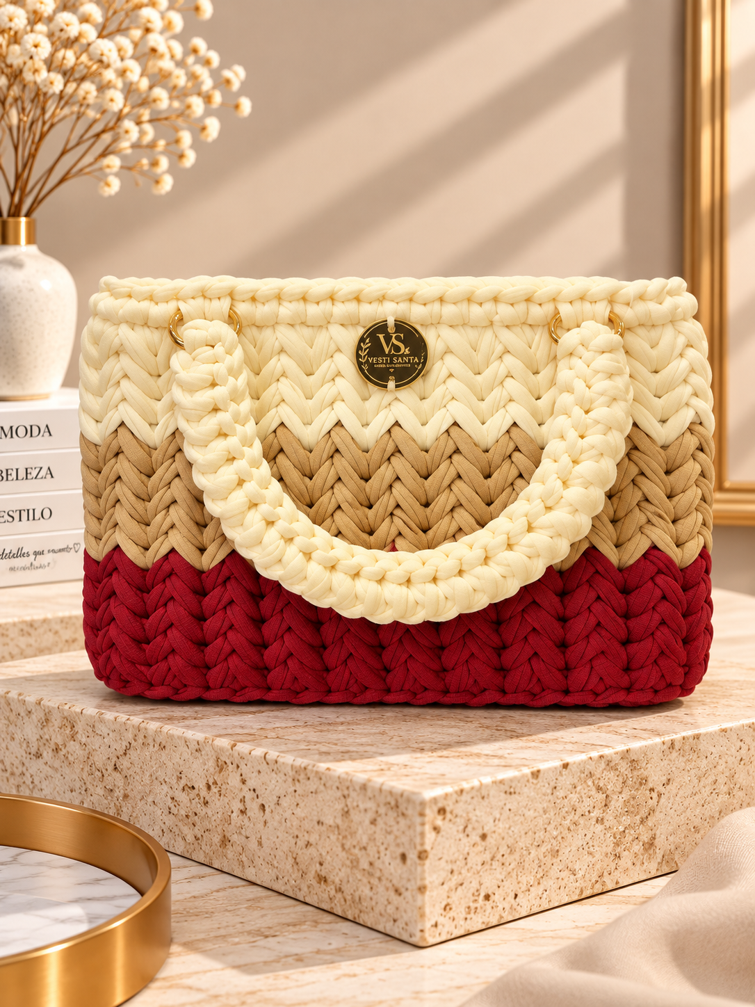 Mala trançada em creme, castanho e vermelho com alças grossas e placa dourada, sobre base de pedra cremosa.