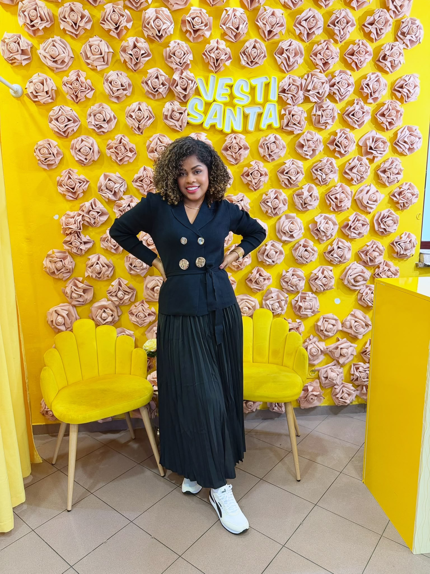 Mulher com casaco preto e saia preta numa loja com decoração amarela e flores cor de rosa na parede.