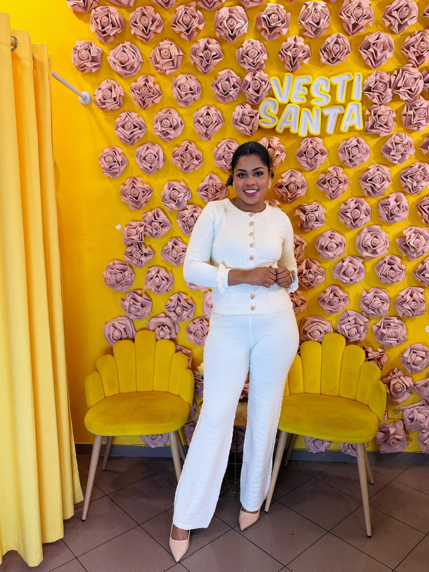 Mulher com conjunto branco em ambiente decorado com flores rosa e parede amarela com texto VESTI SANTA