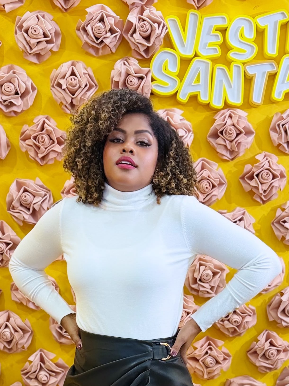 Mulher com camisola branca de gola alta e saia preta em fundo amarelo com flores cor-de-rosa e texto VESTI SANTA