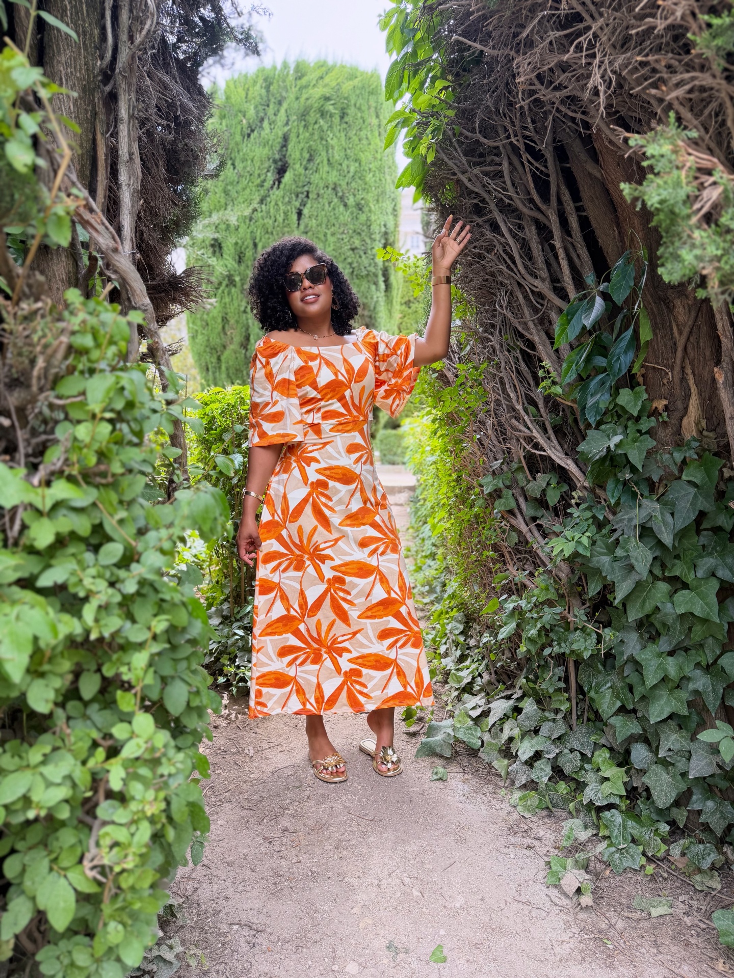 Mulher com vestido floral laranja e branco e óculos escuros num caminho com vegetação