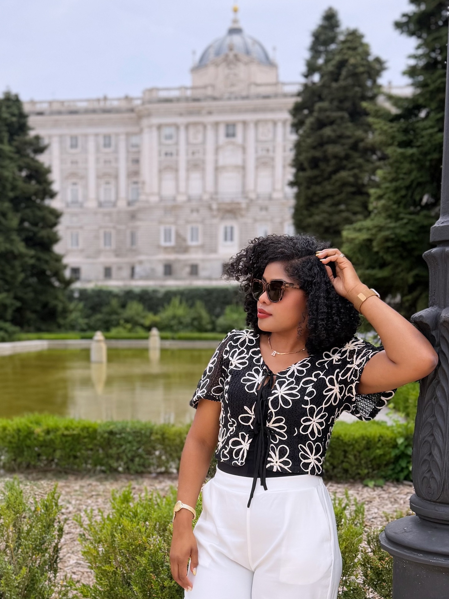 Mulher com top preto com flores brancas, calças brancas, óculos de sol castanhos, pulseira dourada, junto a um lampião em parque com palácio ao fundo