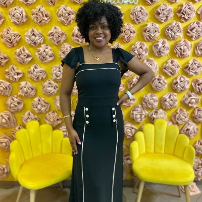 Mulher em vestido preto com detalhes brancos diante de fundo amarelo com rosas 3D rosa e cadeiras amarelas.