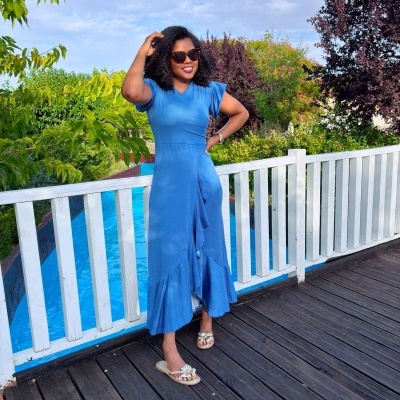 Mulher com vestido azul comprido com folho, óculos de sol e sandálias douradas de pé em passadiço de madeira junto a piscina e vegetação