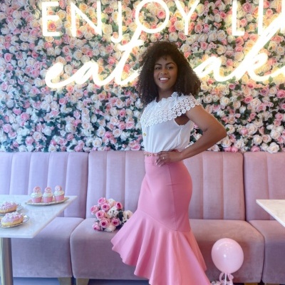 Mulher com blusa branca e saia rosa posando em ambiente decorado com flores artificiais.