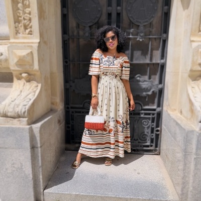 Mulher com vestido longo floral e riscas, mala trançada e sandálias douradas