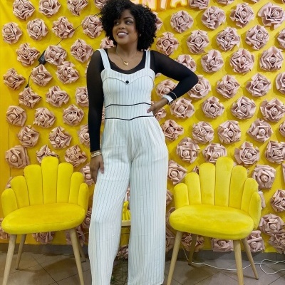 Mulher com macacão branco de riscas sob blusa preta, cadeiras amarelas, parede amarela com flores cor de rosa e texto VESTISANTA