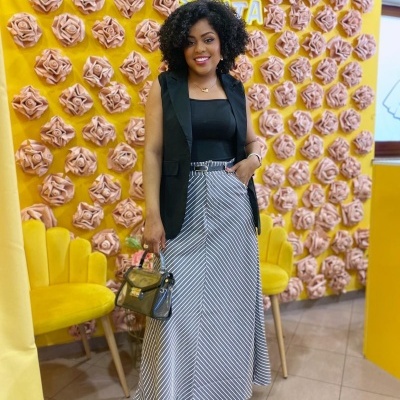 Mulher com colete preto e saia longa preta e branca em fundo amarelo com flores e cadeiras amarelas