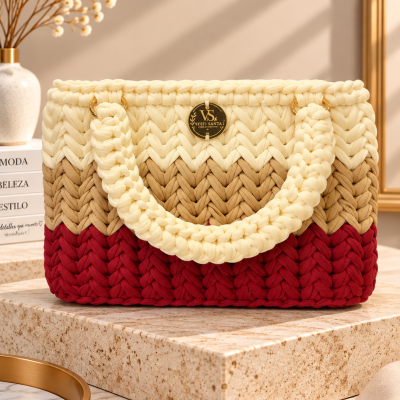 Mala trançada em creme, castanho e vermelho com alças grossas e placa dourada, sobre base de pedra cremosa.