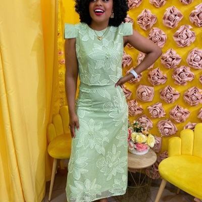Mulher com vestido verde água florido em ambiente amarelo com flores de parede e cadeiras amarelas.