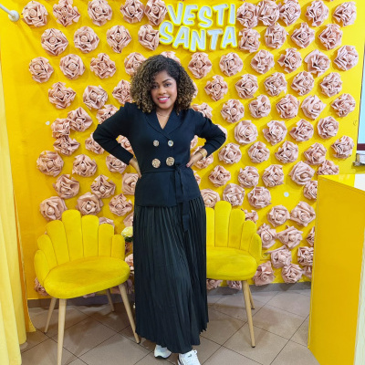 Mulher com casaco preto e saia preta numa loja com decoração amarela e flores cor de rosa na parede.