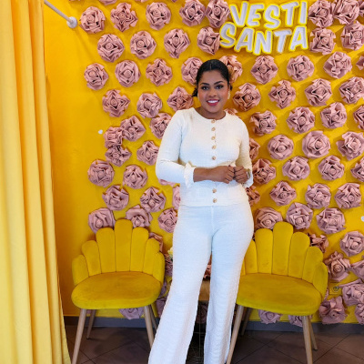 Mulher com conjunto branco em ambiente decorado com flores rosa e parede amarela com texto VESTI SANTA