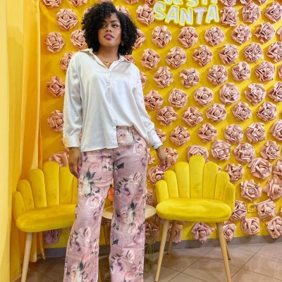 Mulher com camisa branca e calças floridas cor-de-rosa em frente a parede amarela com flores cor-de-rosa e texto VESTI SANTA