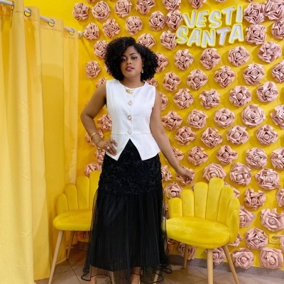 Mulher com colete branco e saia preta longa em parede amarela com flores rosa e cadeiras amarelas